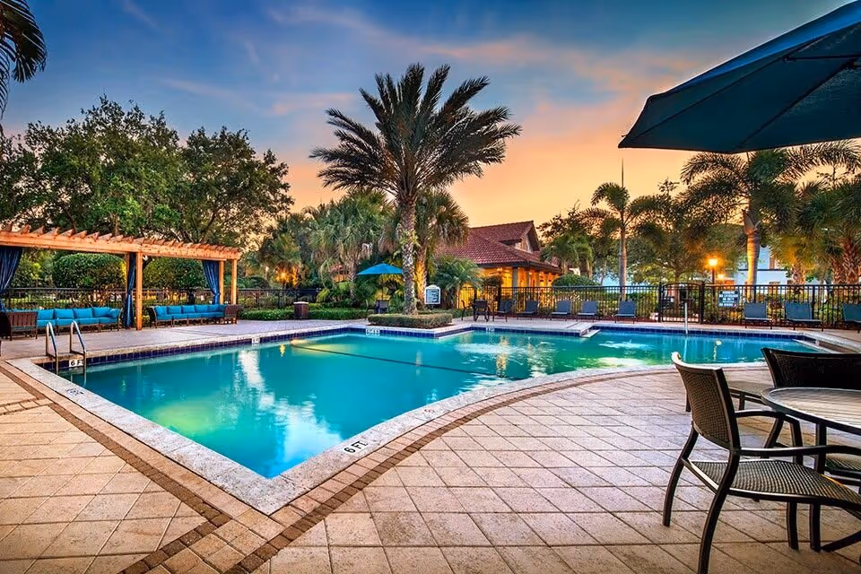 Outdoor swimming pool area with loungers, tables, umbrellas and palm trees at sunset.