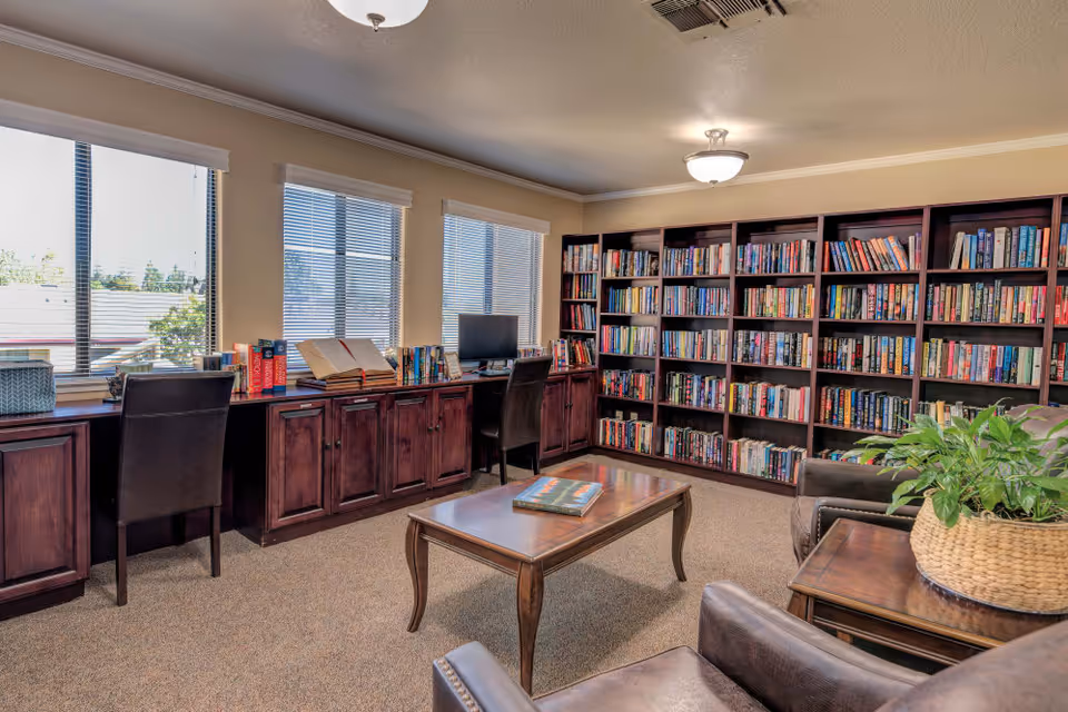 A cozy library room with large windows letting in natural light. The room features dark wooden bookshelves filled with books along one wall, a wooden table in the center with a book on it, and two leather armchairs. There is a long wooden desk under the windows with two chairs and a computer monitor. A potted plant sits on a side table next to one of the armchairs.
