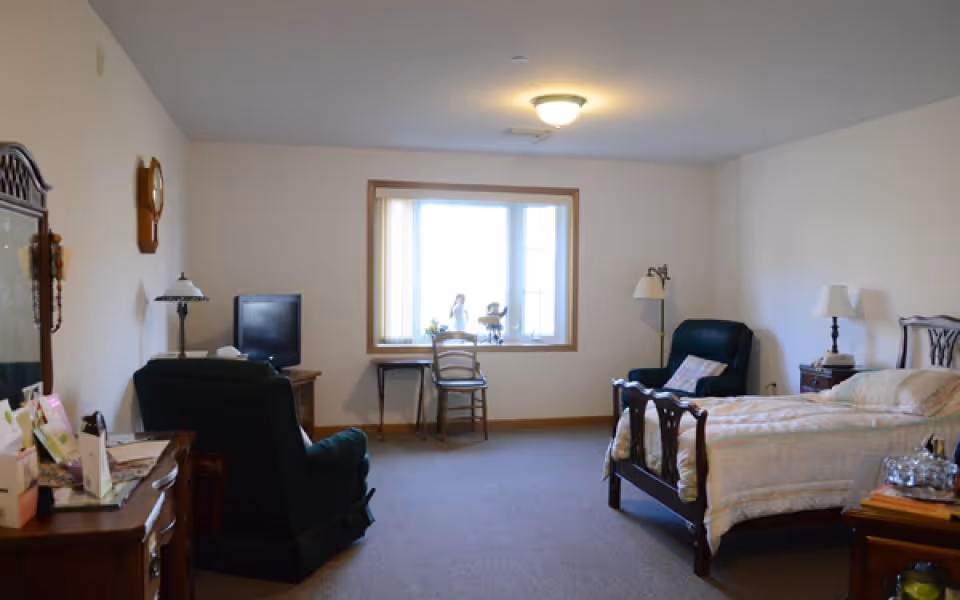 A well-lit bedroom in a senior living facility with a single bed, two armchairs, a wooden dresser with a mirror, a small desk with a chair in front of a large window, and various lamps providing lighting.