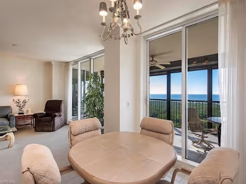 A bright living room with a round wooden table surrounded by four cushioned chairs in the foreground. Behind the table, there is a sliding glass door leading to a balcony with outdoor chairs and a table, overlooking a scenic view of the ocean. The room also features a comfortable armchair, a side table with a lamp, and a chandelier hanging from the ceiling.