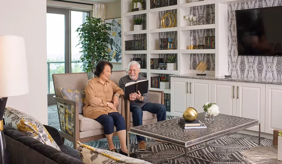 An elderly couple sitting in armchairs in a well-decorated living room area. The man is holding an open book and smiling while the woman looks on with a smile. The room features built-in white shelves filled with books and decorative items, a large flat-screen TV mounted on the wall, a patterned rug, a modern coffee table with decorative objects, and a large window with a view of greenery outside.