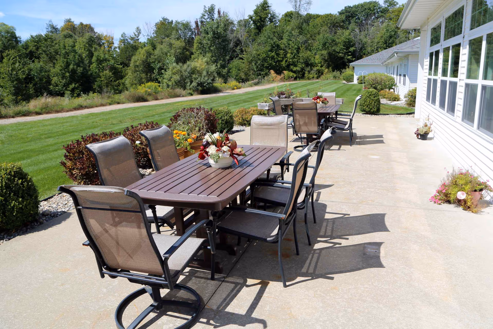 Outdoor concrete patio with multiple dining tables and chairs alongside a white building overlooking a landscaped lawn and trees.