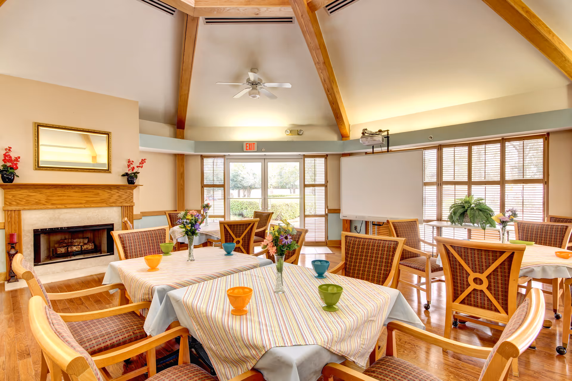 A bright and spacious common area with several tables covered in striped tablecloths, each adorned with colorful bowls and vases of flowers. The room features wooden chairs with patterned cushions, a fireplace with a mirror above it, large windows with blinds letting in natural light, and a ceiling fan with exposed wooden beams. A whiteboard and projector are also visible near the windows.