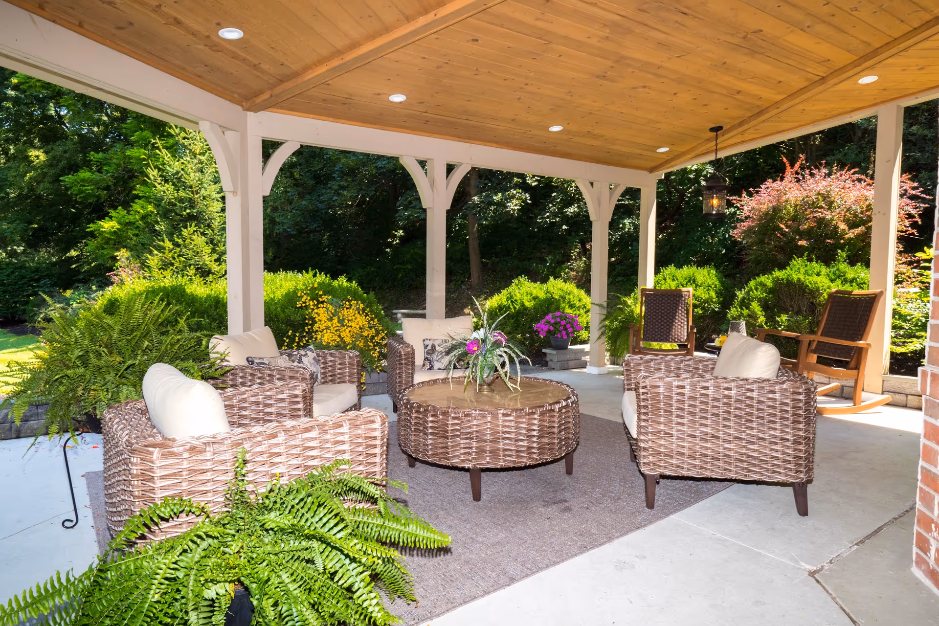 Covered outdoor seating area with wicker chairs and a round wicker table on a rug, surrounded by lush green plants and trees in the background.