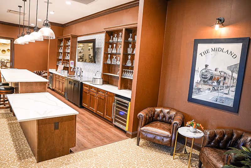Communal kitchen and seating area with marble-topped islands, wood cabinetry and glassware shelves, pendant lights, and leather armchairs beneath framed artwork.