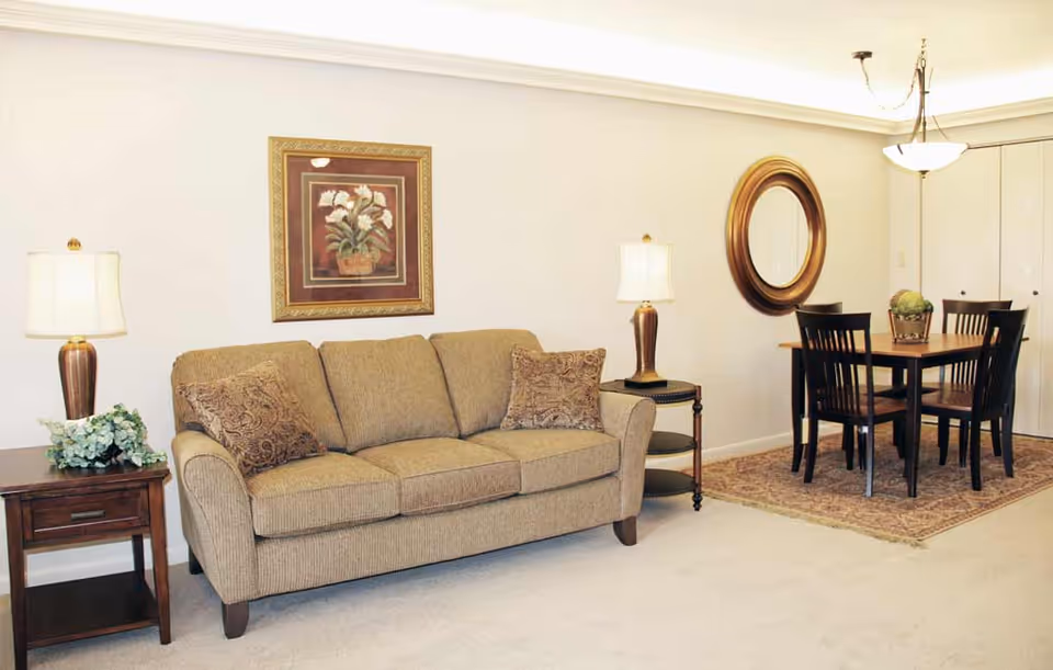 Beige living room with a three-seat sofa, side tables and lamps, wall art, and a dining table with chairs in the background.
