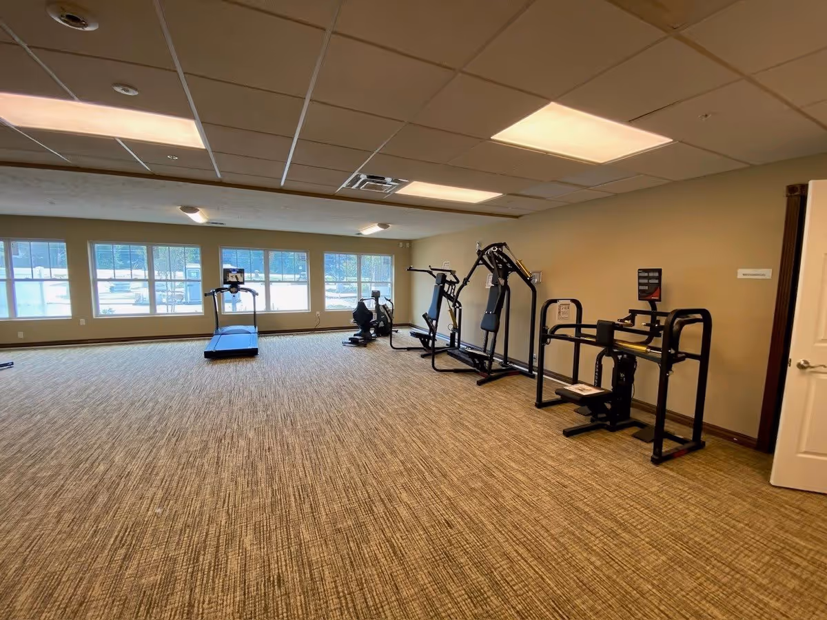 A spacious exercise room with large windows letting in natural light. The room has carpeted flooring and beige walls. Along one wall, there are several pieces of exercise equipment including a treadmill, an elliptical machine, and various strength training machines.
