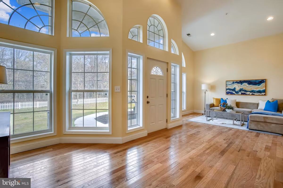 Bright living room area with large arched and rectangular windows letting in natural light. The room features light-colored walls, hardwood floors, a beige sectional sofa with blue and yellow pillows, a floor lamp, a coffee table, and a piece of abstract wall art above the sofa. The front door is white with glass panels on either side.