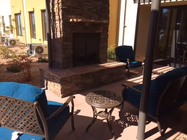 Outdoor patio area with a stone fireplace surrounded by metal chairs with blue cushions and a small round metal table. The patio is adjacent to a building with yellow walls and several windows, and there are air conditioning units visible in the background.