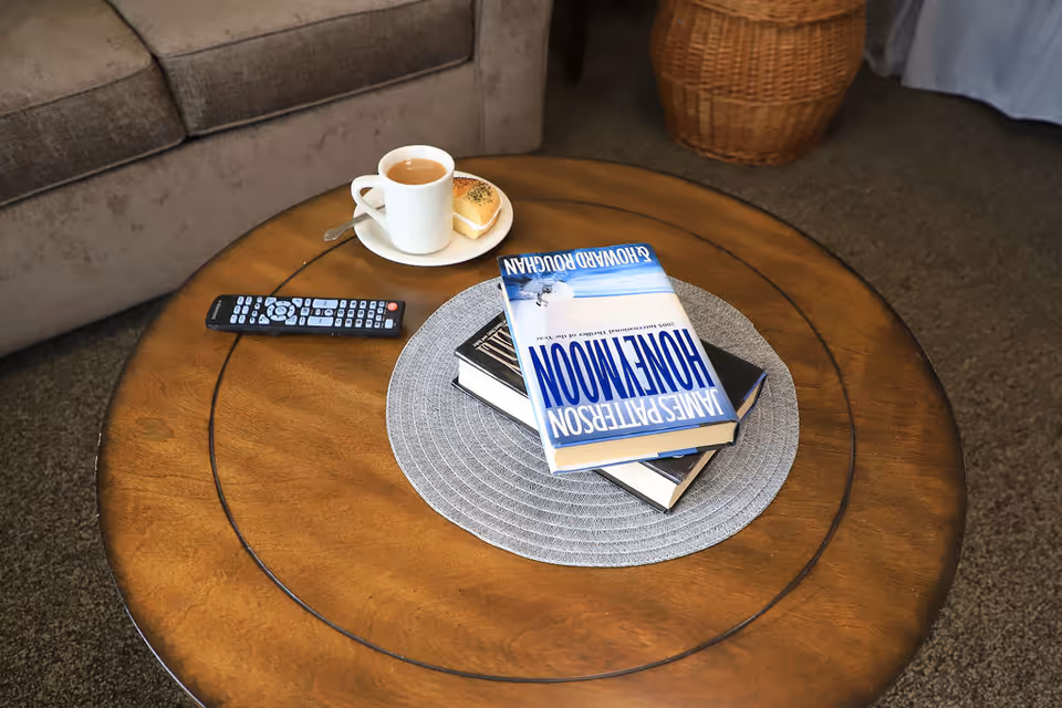 A round wooden coffee table with a gray woven placemat holding three stacked books, a TV remote control, and a white cup of coffee on a saucer with a small pastry. A gray sofa and a wicker basket are visible in the background.
