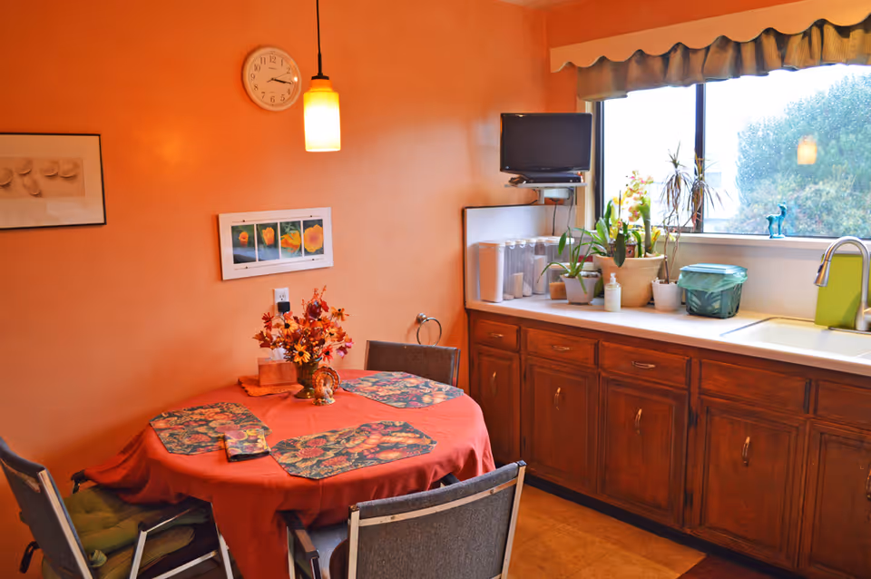 Small dining area and kitchenette with a round table covered in a red tablecloth, pendant light and wall clock, wooden cabinets and a countertop with plants and a sink under a window.