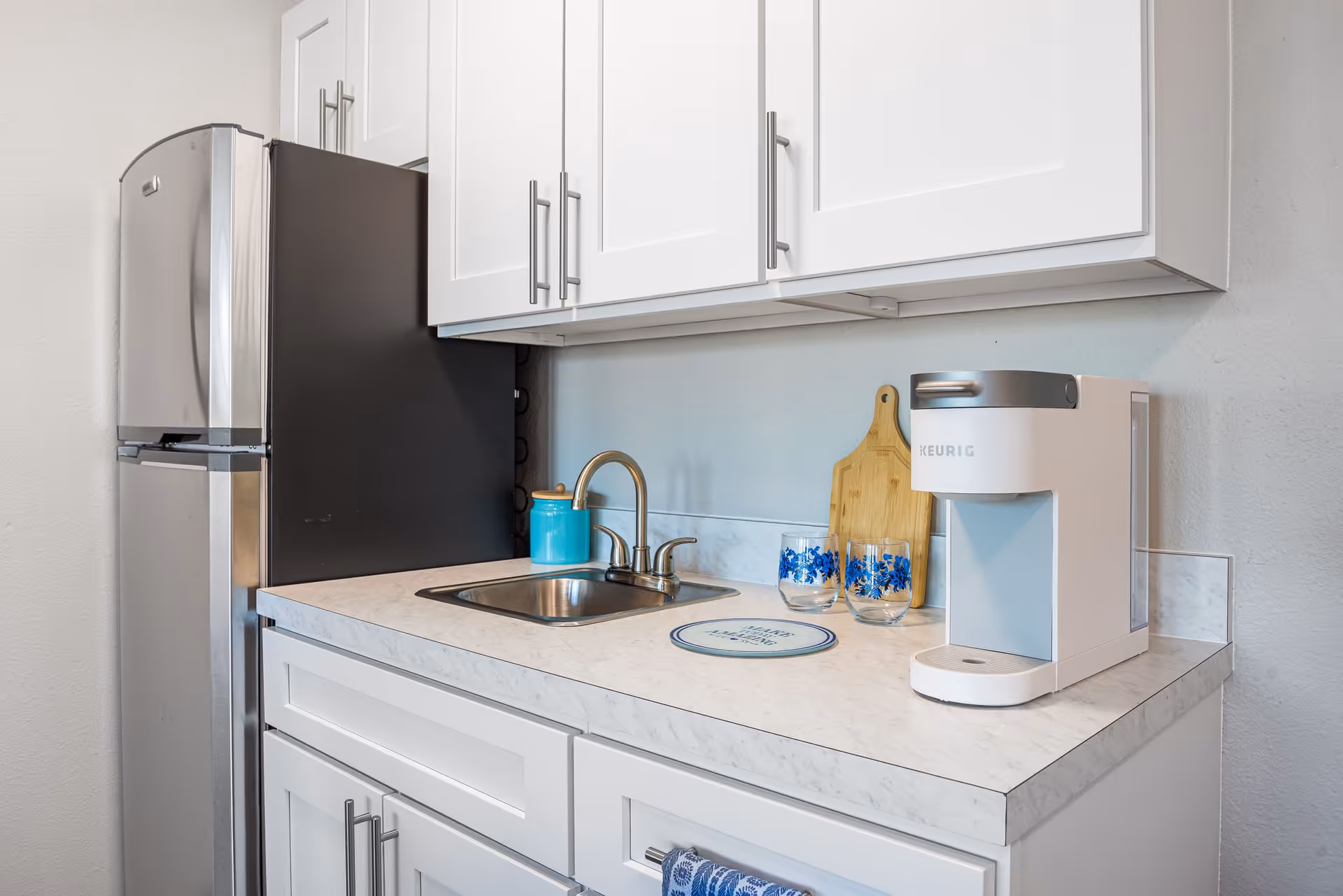 A compact kitchen area featuring a stainless steel refrigerator, white cabinets with silver handles, a small sink with a faucet, a white Keurig coffee maker, two glasses with blue floral designs, a wooden cutting board, and a blue container on the countertop.
