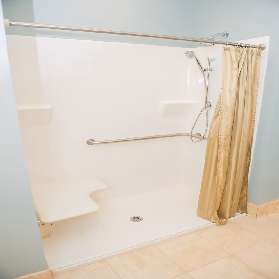 Walk-in accessible shower with a fold-down seat, horizontal grab bar, handheld showerhead, and a beige shower curtain.