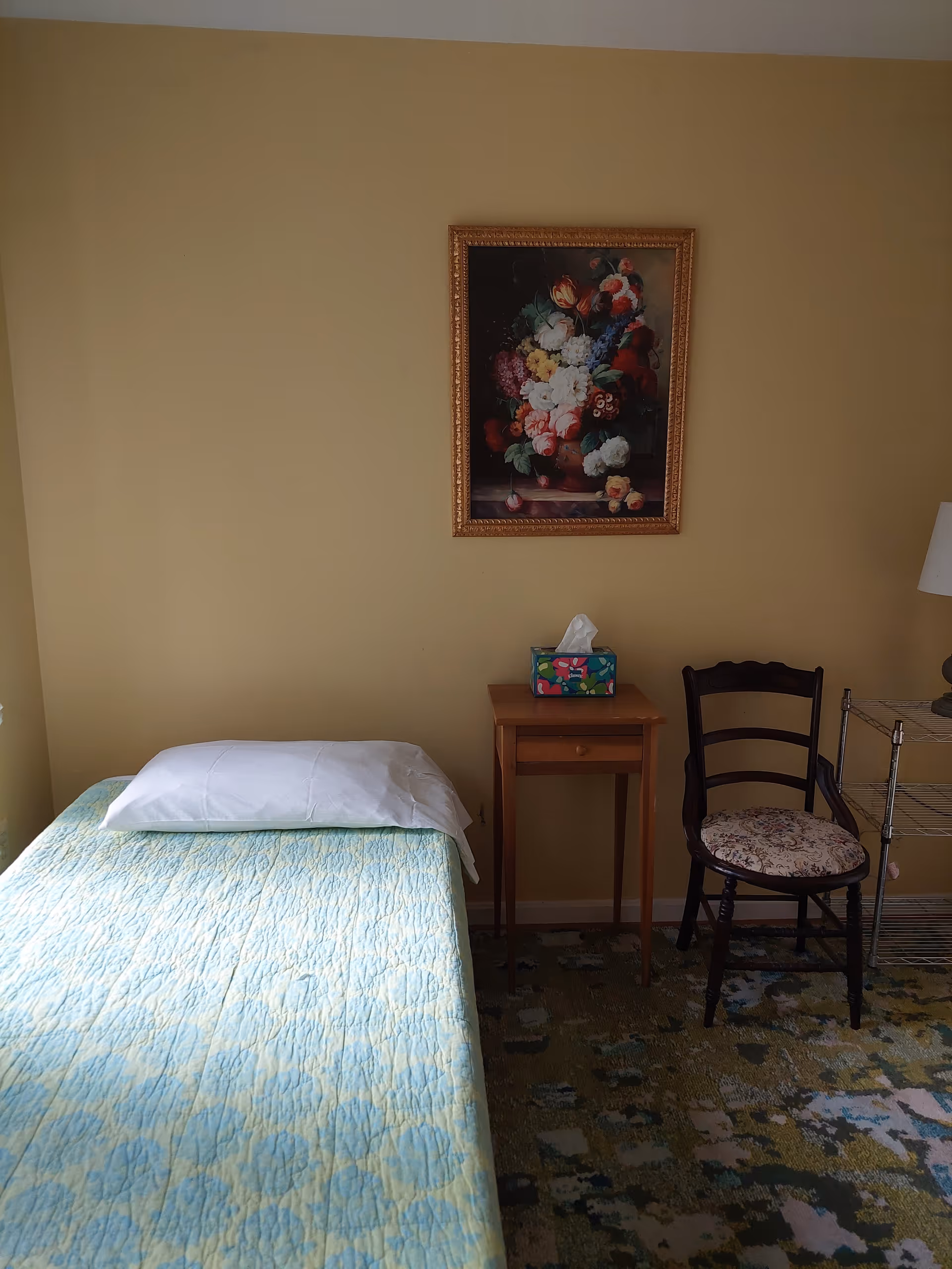A small bedroom with a single bed covered with a light blue and green patterned quilt and a white pillow. Next to the bed is a wooden nightstand with a colorful tissue box on top. A wooden chair with a floral cushion is placed beside the nightstand. A framed painting of a floral bouquet hangs on the beige wall above the nightstand. The floor has a patterned carpet with various shades of green and blue.