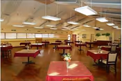 Spacious dining room with multiple tables covered in red tablecloths, each with a small floral centerpiece. The room has wooden flooring, large windows along one wall, and modern rectangular ceiling lights. Chairs are arranged around the tables, and the walls are decorated with framed pictures and plants.