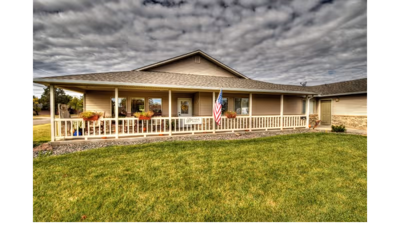 Exterior view of a single-story building with beige siding and a covered porch. The porch has white railings, several potted plants, and an American flag displayed near the entrance. The building is surrounded by a well-maintained green lawn under a cloudy sky.