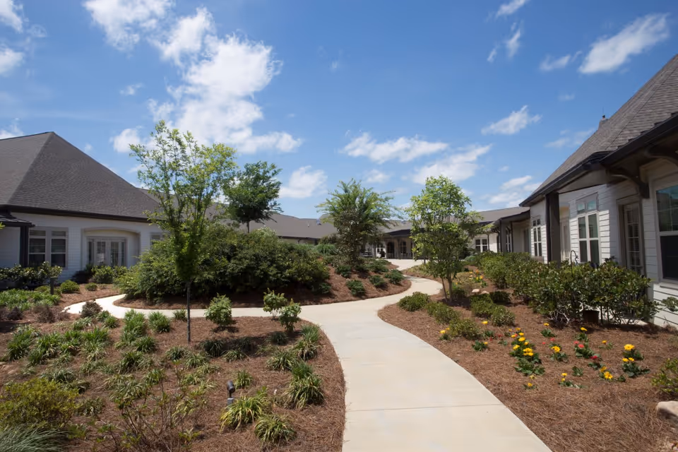 Curving paved walkway winds through a landscaped courtyard with shrubs, flowers, and small trees between single-story building wings under a blue sky.
