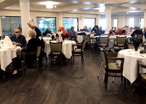A bright dining room with round tables covered in white tablecloths and many seniors seated and dining.
