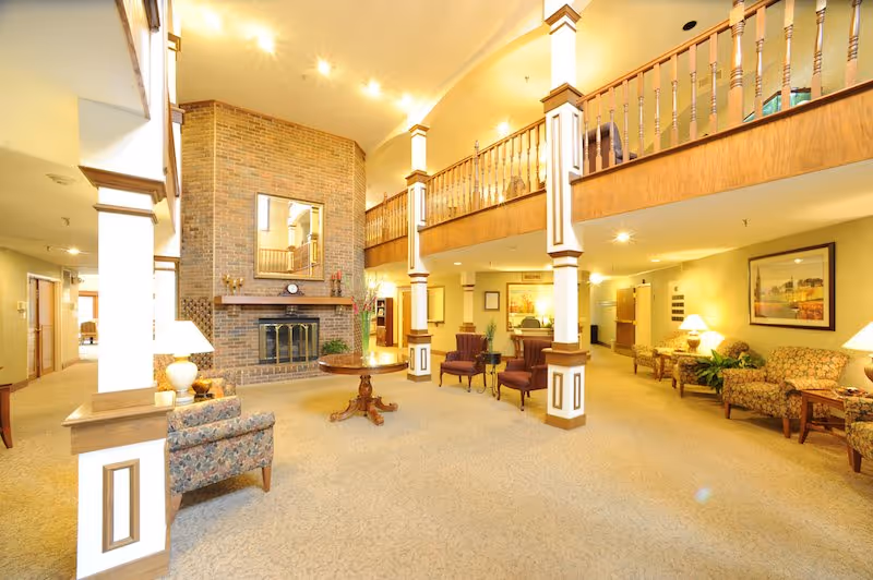 Spacious and well-lit senior living facility common area with a high ceiling, large brick fireplace, wooden railing on the upper level, multiple seating arrangements including armchairs and sofas, decorative lamps, and framed artwork on the walls.