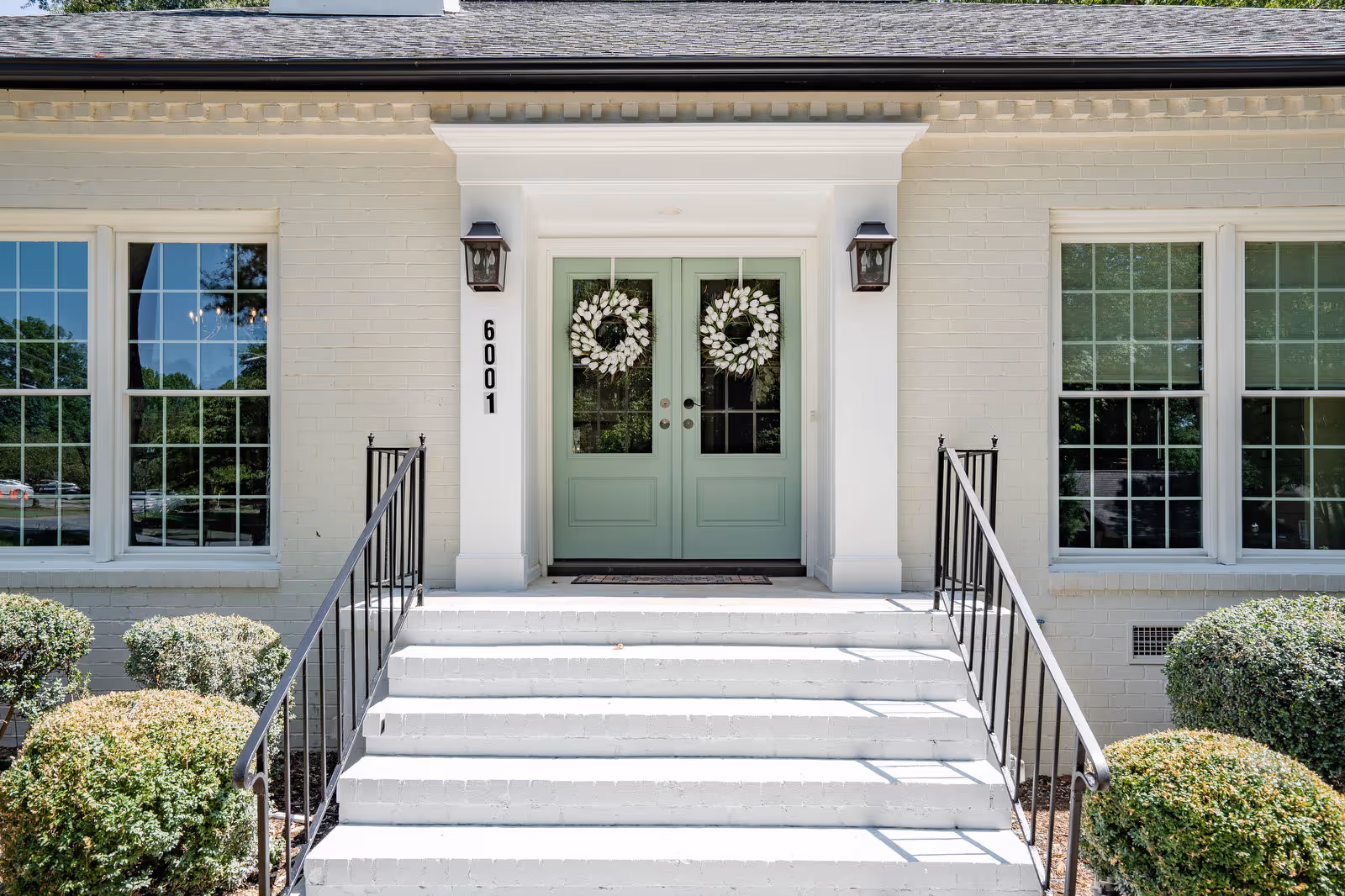 Front entrance of a building with white brick exterior, green double doors decorated with white wreaths, black metal railings on either side of white steps leading up to the doors, and large windows on both sides of the entrance.