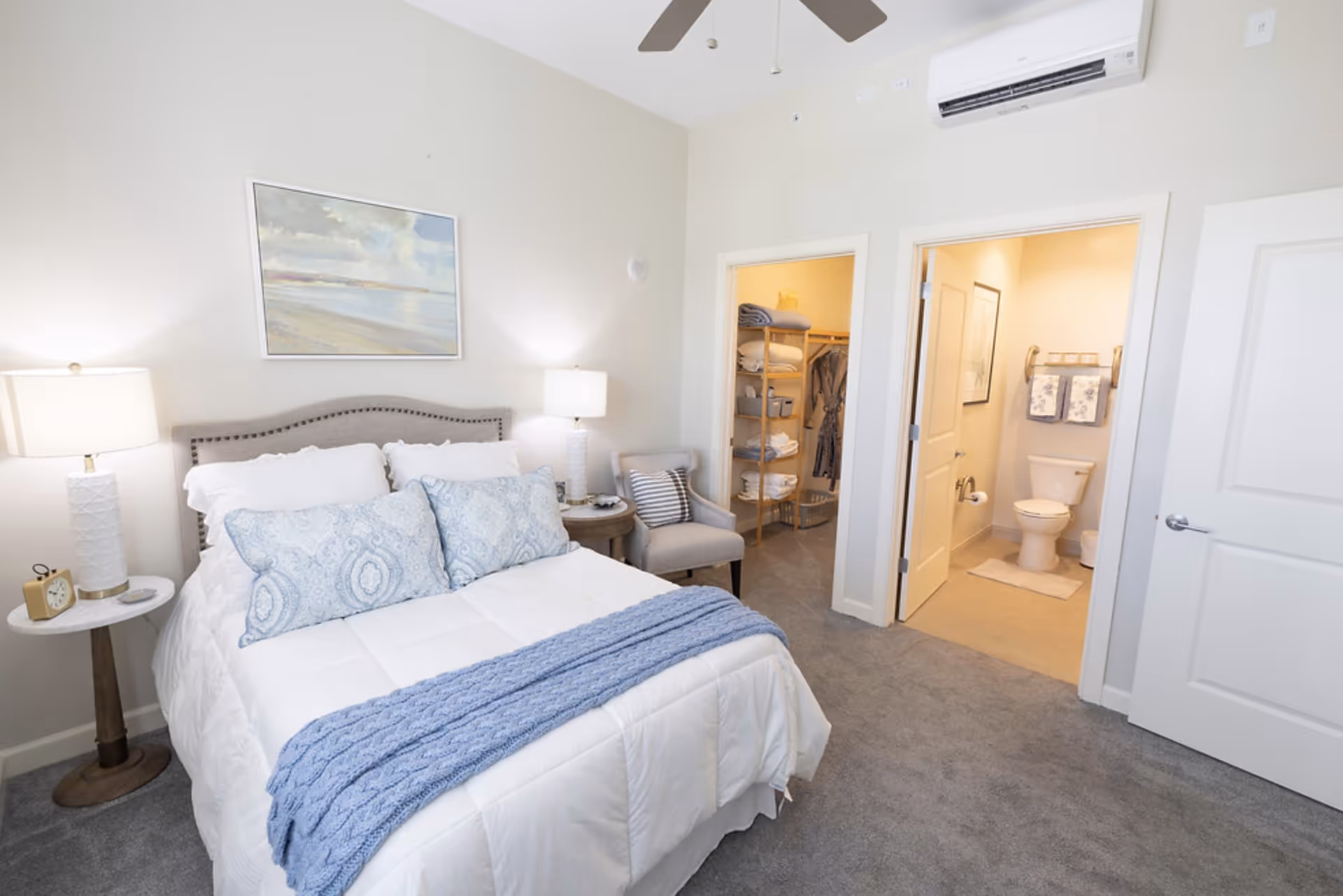A cozy bedroom with a neatly made bed featuring white bedding and blue patterned pillows and a blue throw blanket. Two bedside tables with white lamps flank the bed. A framed beach scene hangs above the bed. To the right, there is an open door leading to a bathroom with a toilet and towels, and another open door revealing a walk-in closet with shelves holding folded towels and hanging clothes. The room has light-colored walls, gray carpet, a ceiling fan, and an air conditioning unit mounted on the wall.