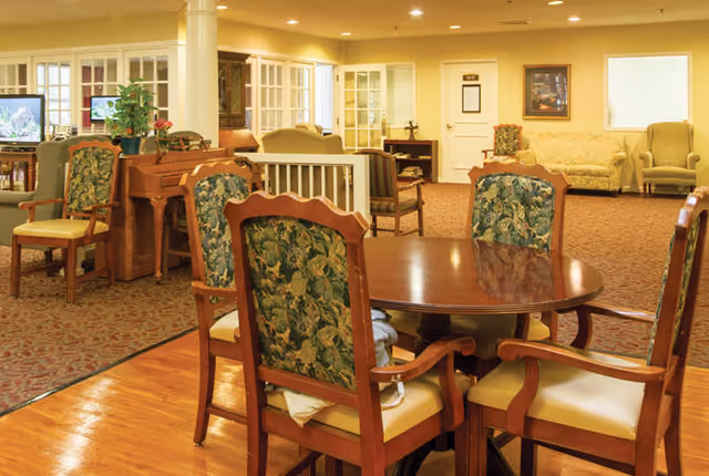 A spacious senior living common area with wooden chairs featuring floral upholstery around a polished wooden table. The room has a warm yellow lighting, carpeted and wooden floors, a piano, a TV, and several sofas and armchairs arranged for socializing.