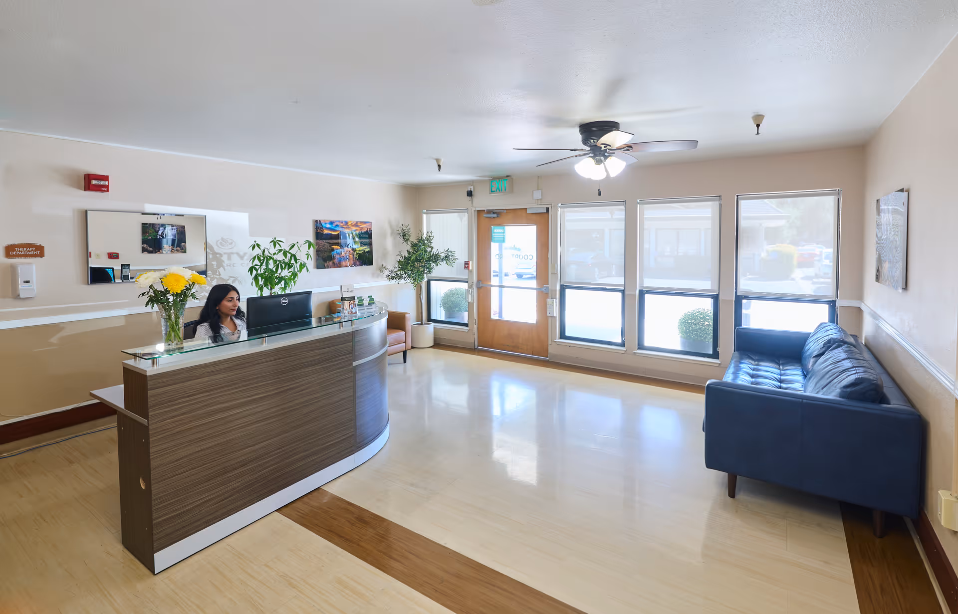 Reception area of Courtyard Healthcare Center with a woman sitting behind a curved wooden reception desk with a computer. The room has large windows with blinds, a ceiling fan with lights, a blue couch along the right wall, and plants near the entrance door.