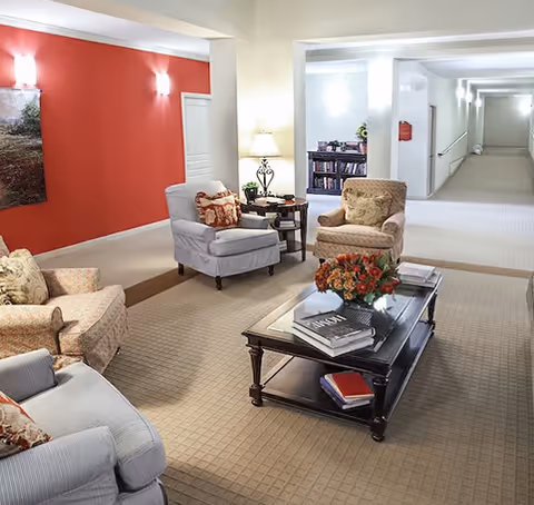 A cozy seating area in a senior living facility with four upholstered armchairs arranged around a dark wooden coffee table that holds books and a floral arrangement. The room features a red accent wall with a landscape painting, soft lighting from wall sconces and a table lamp, and a carpeted floor. In the background, there is a hallway and a bookshelf filled with books.