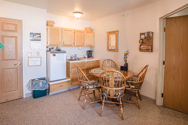 A small kitchen and dining area in a senior living facility with wooden cabinets, a white mini refrigerator, a coffee maker, and a round wooden table with four wooden chairs. The walls are light-colored, and there is a small mirror and a decorative wall hanging. Two wooden doors are visible, one on the left and one on the right.
