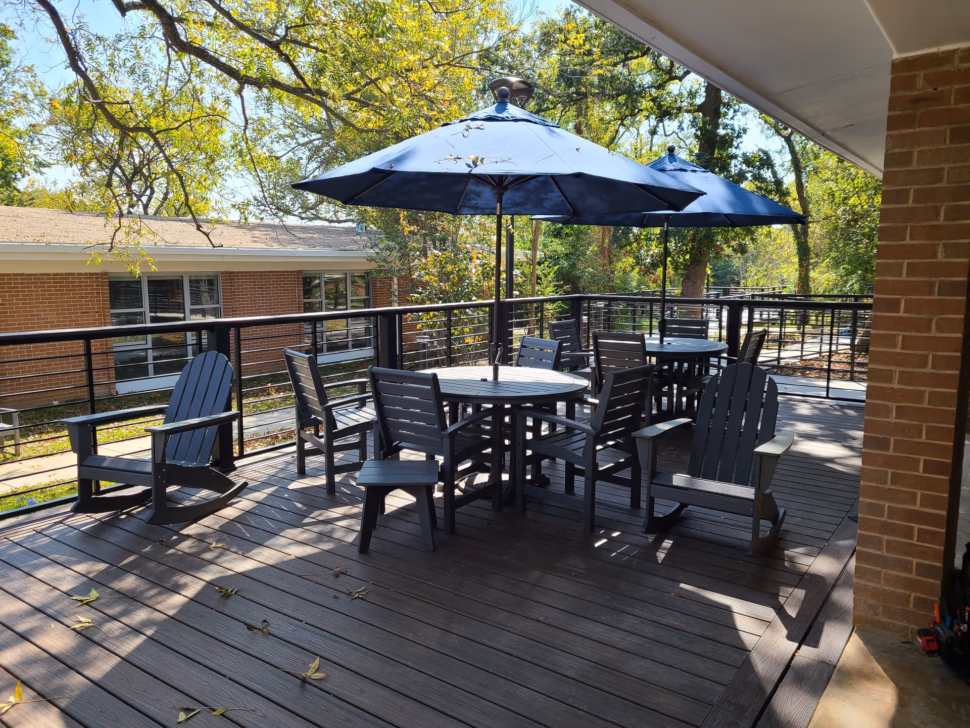 Outdoor patio area with wooden deck flooring, featuring two round tables each with a blue umbrella and surrounded by multiple dark wooden chairs and rocking chairs. The patio is bordered by a black metal railing and is adjacent to a brick building. Trees and greenery are visible in the background under a clear sky.