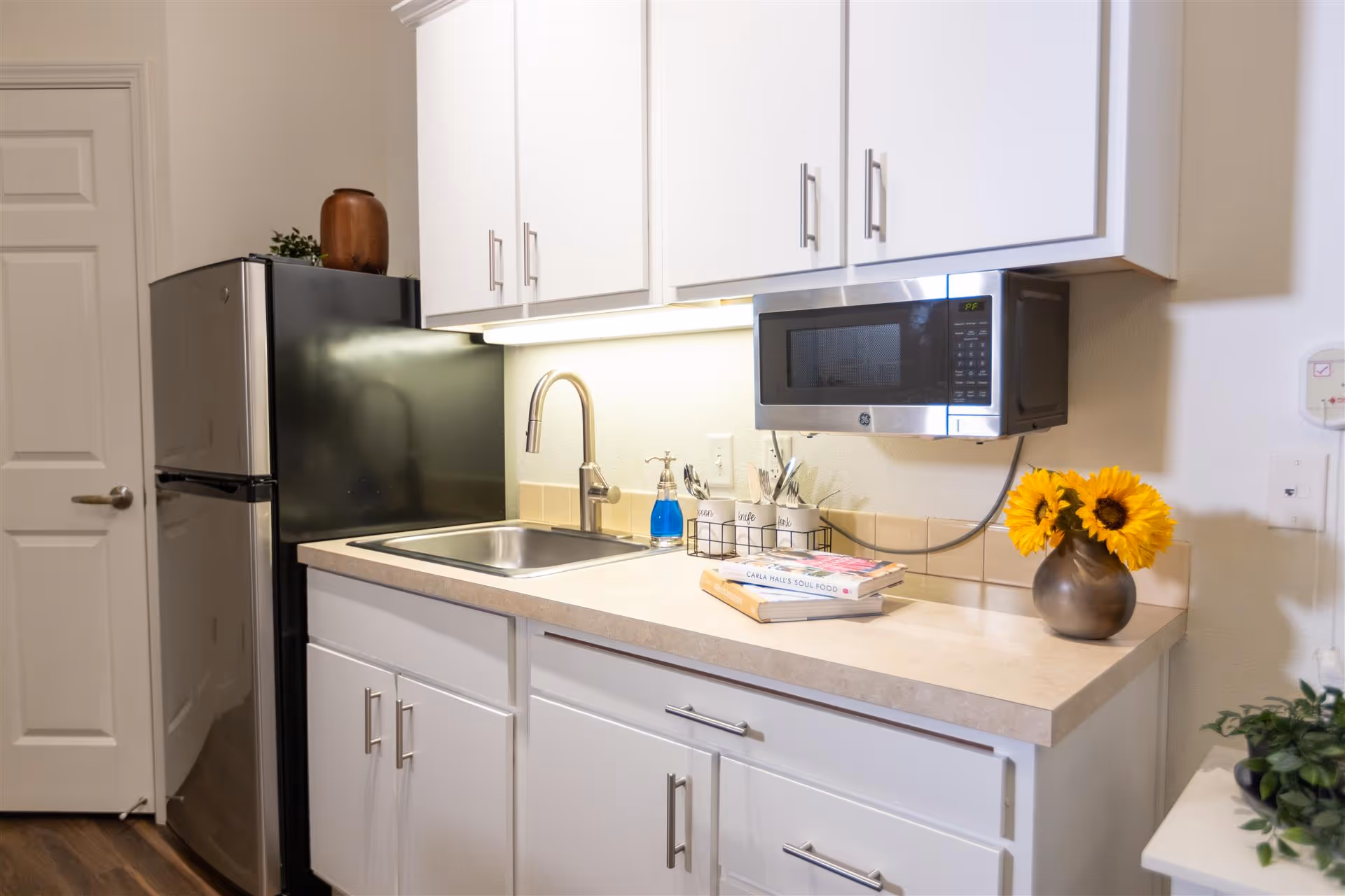 A clean and modern kitchen area with white cabinets, a stainless steel refrigerator, a sink with a faucet, a mounted microwave, a vase with sunflowers, and some books and kitchen utensils on the countertop.