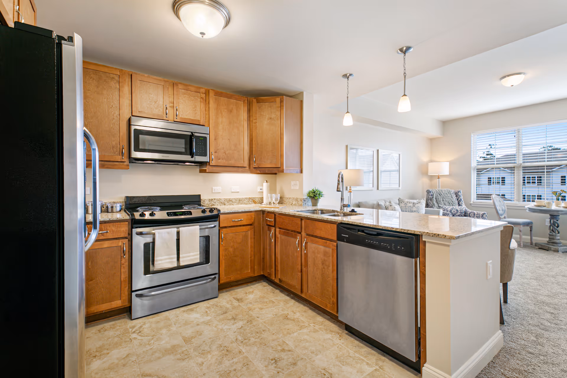 Open kitchen with stainless steel refrigerator, oven and dishwasher, wooden cabinets and a granite-topped island overlooking a living area.