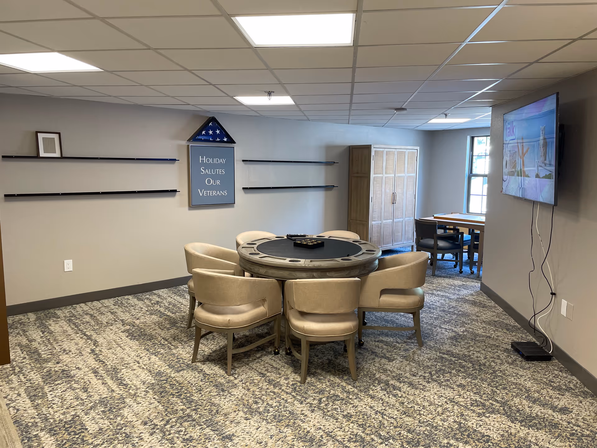 Communal activity room with a round game table surrounded by chairs, a wall-mounted TV, shelving, and a sign honoring veterans.
