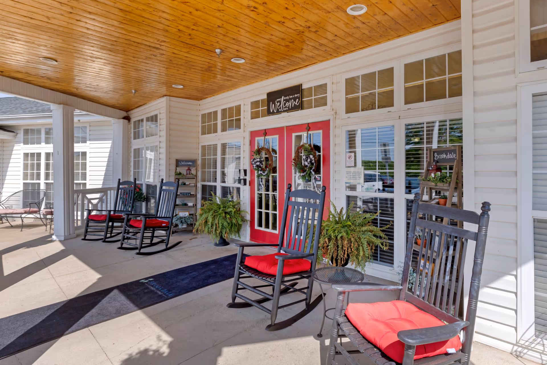 Covered outdoor porch area with black rocking chairs featuring red cushions, small tables, potted plants, and double red doors with wreaths. The porch has a wooden ceiling and white siding walls with large windows.