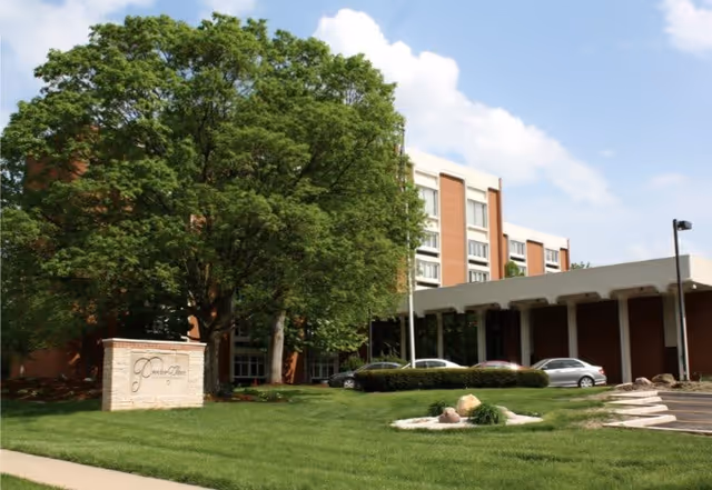 Exterior view of Proctor Place Senior Living facility showing a multi-story brick building partially obscured by large green trees, a manicured lawn, a stone sign with the facility name, several parked cars, and a clear blue sky with some clouds.