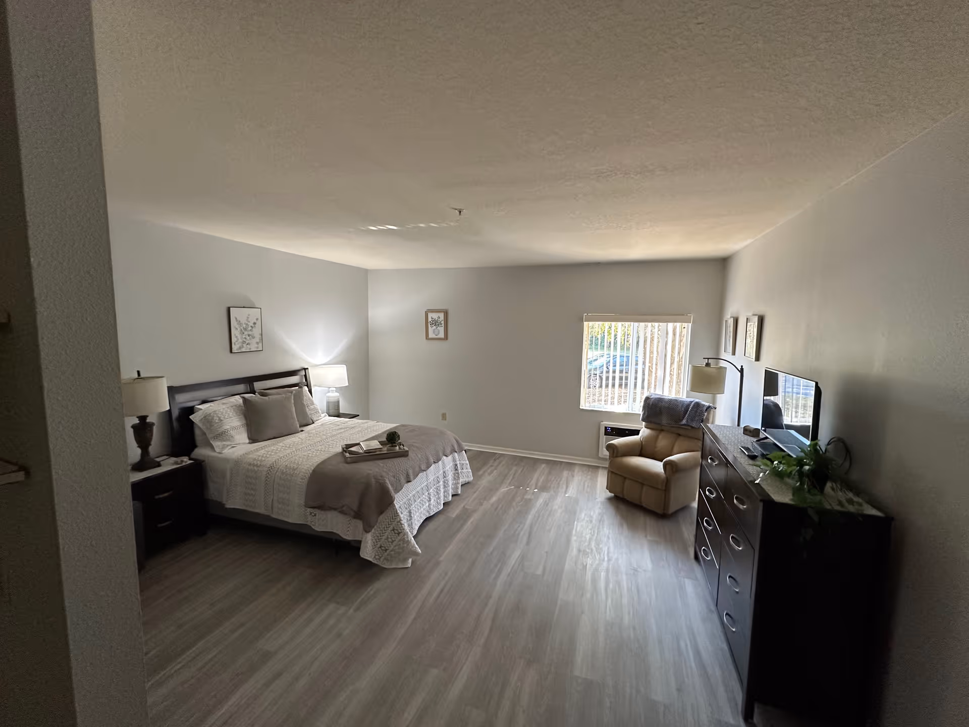 A spacious senior living bedroom with a large bed dressed in white and beige bedding, two bedside tables with lamps, a comfortable armchair near a window with vertical blinds, a dark wooden dresser with a TV on top, and light-colored wood flooring.
