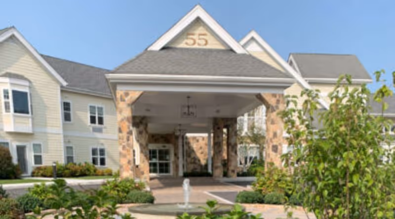 Front entrance of a senior living building with a covered porte-cochere supported by stone columns, a small fountain, and the number '55' displayed above.