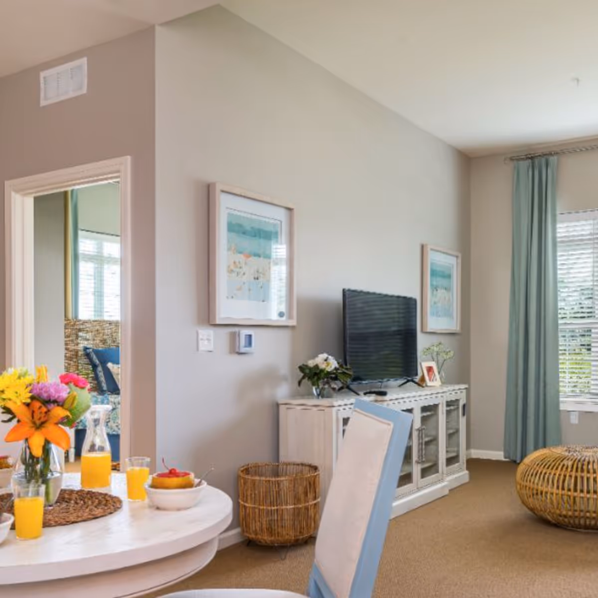 A bright living room with beige walls and carpet, featuring a white TV stand with a flat-screen TV and decorative items. Two framed artworks hang on the wall above the TV stand. A round white dining table with a vase of colorful flowers, glasses of orange juice, and a bowl of fruit is partially visible in the foreground. A wicker basket and a round wicker ottoman are also in the room. Light blue curtains frame a window with blinds, and an open doorway leads to another room with a blue chair and a window.