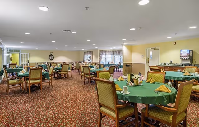 A spacious dining room with multiple round tables covered with green tablecloths and set with yellow folded napkins, cups, and small centerpieces. The room has patterned carpet flooring, beige walls, and ceiling lights. There is a beverage dispenser and a counter area on the right side, and windows with curtains in the background.