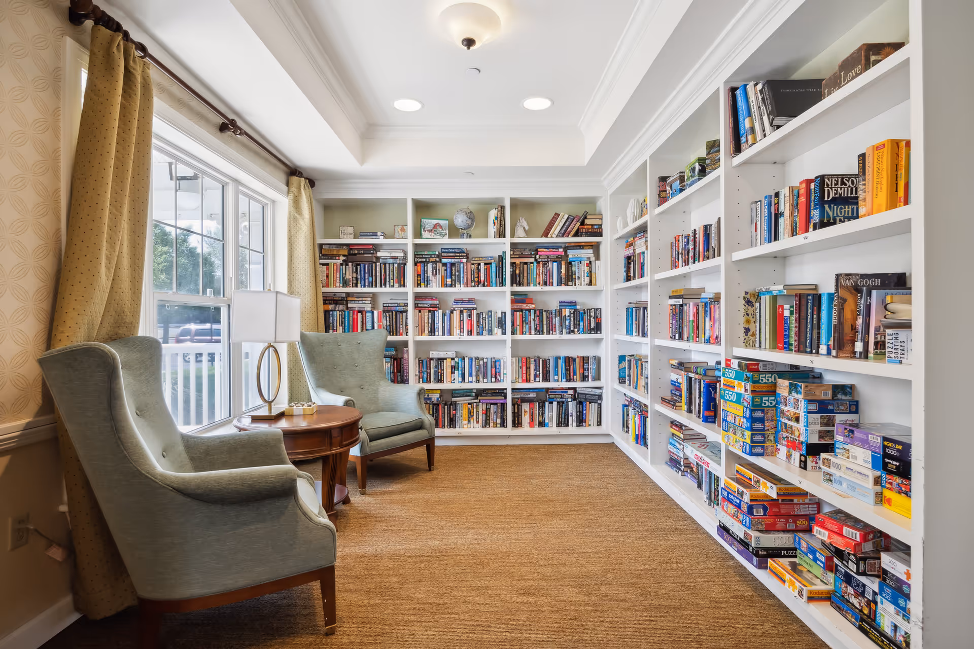 A cozy reading room with two light green armchairs positioned around a small wooden round table with a lamp. The room features large windows with yellow curtains letting in natural light. The walls are lined with white bookshelves filled with books and puzzles. The floor is carpeted in a warm brown tone.