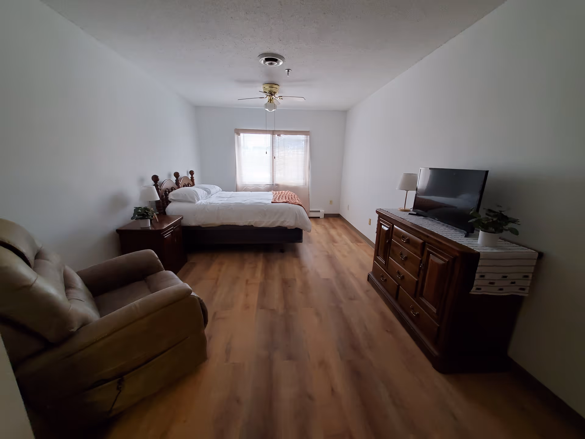 Simple furnished bedroom with a bed, recliner, dresser with a TV and a window at the far end.