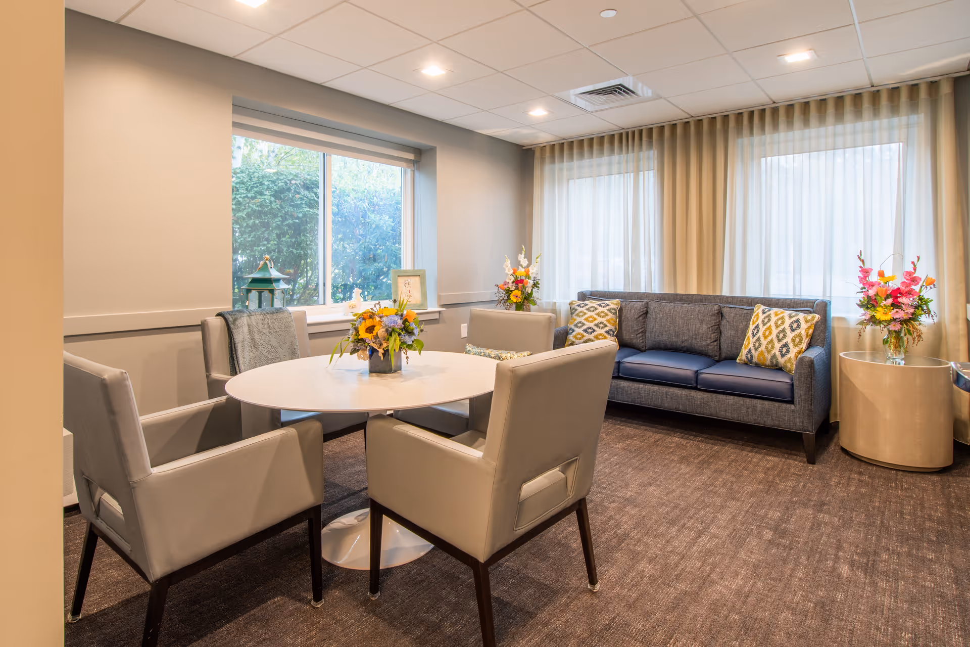 A cozy sitting area in a senior living facility with a round white table surrounded by four beige armchairs. Behind the table is a gray sofa with patterned cushions. The room has large windows with sheer curtains allowing natural light to enter. There are flower arrangements on the table and side tables, and a framed picture and decorative lantern on the window sill.