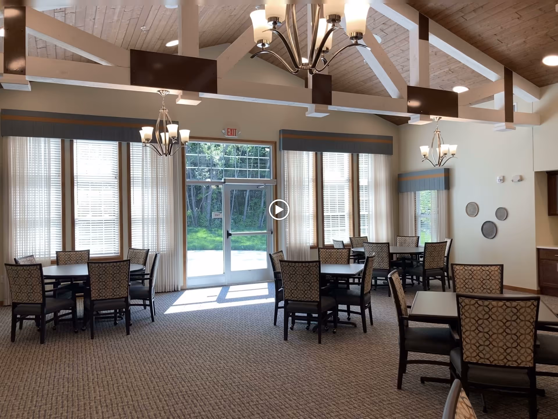 A bright dining room in a senior living facility with multiple tables and chairs arranged neatly. Large windows with sheer curtains allow natural light to fill the room. The ceiling features exposed wooden beams and modern chandeliers. A glass door at the back leads to an outdoor area with greenery visible outside.