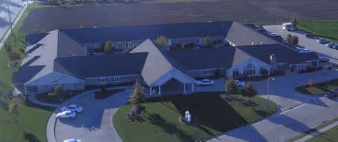 Aerial view of a large single-story senior living facility building with multiple roof sections, surrounded by green lawns and parking areas, under clear daylight.