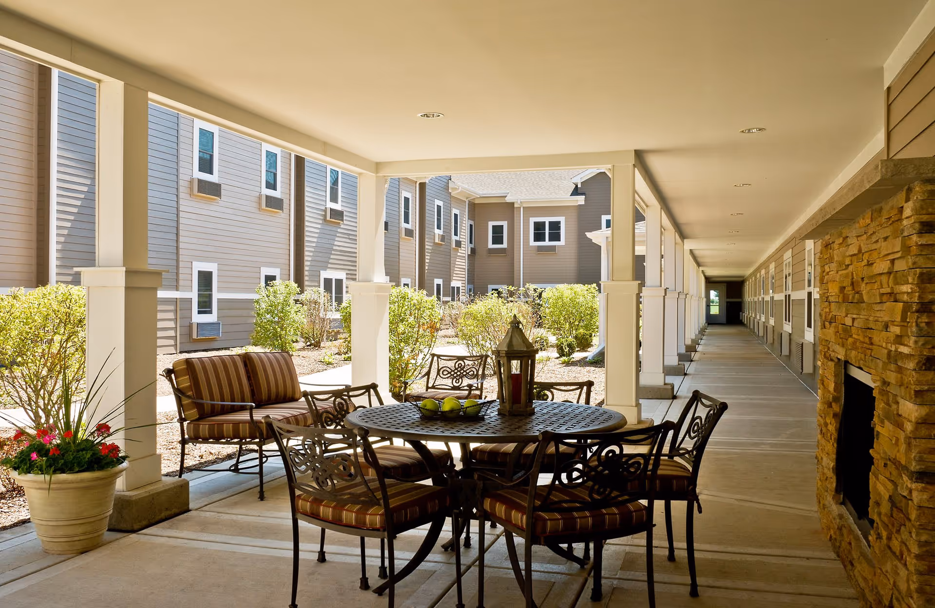 Covered outdoor patio area with a round metal table and six striped cushioned chairs. There is a decorative lantern and a bowl of green apples on the table. The patio is surrounded by beige buildings with multiple windows and small bushes. A stone fireplace is visible on the right side of the patio.