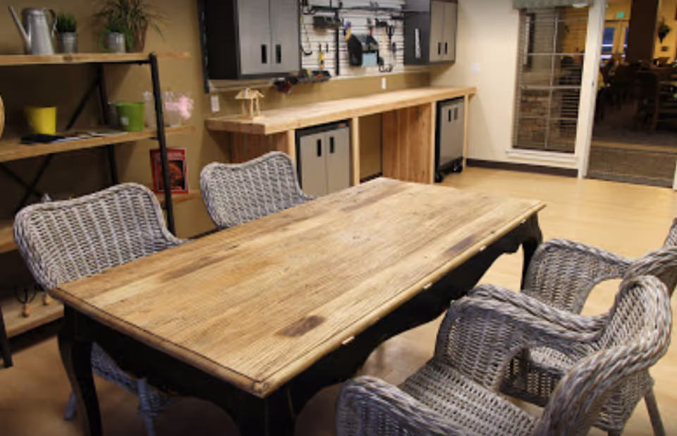 A cozy interior room featuring a wooden table surrounded by four wicker chairs. In the background, there is a workbench with tools hanging on a pegboard and storage cabinets underneath. A shelving unit with decorative items and plants is visible on the left side, and a glass door leads to another room with more seating.