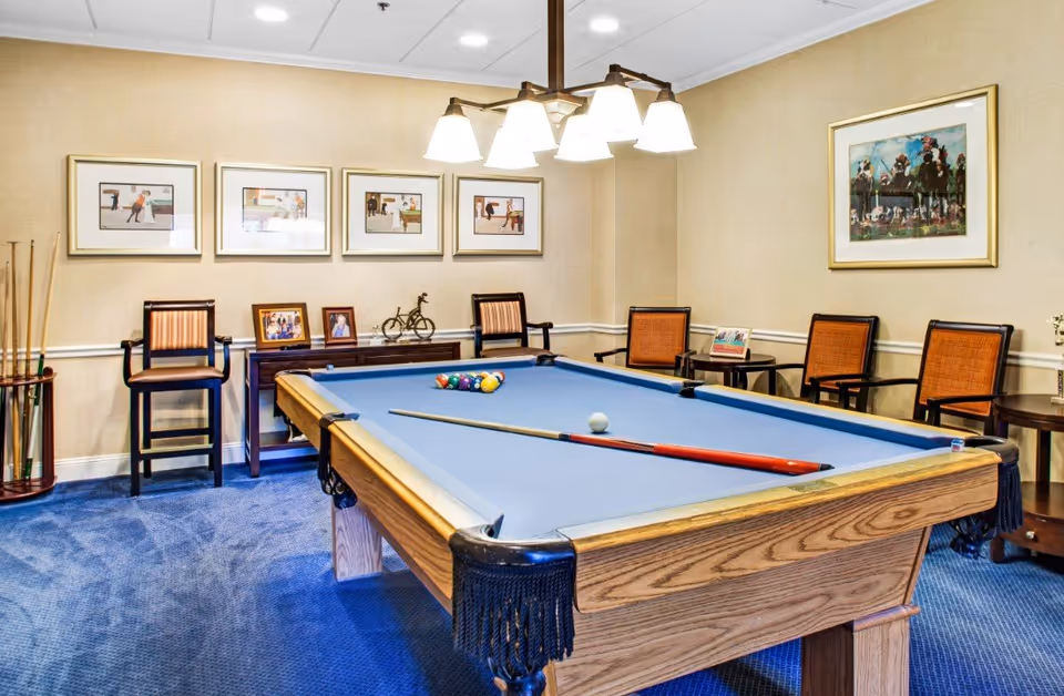 A recreational room with a pool table in the center, featuring a blue felt surface and a cue stick resting on it. The room has blue carpet flooring, beige walls adorned with framed pictures, and several chairs arranged along the walls. A chandelier with multiple lights hangs above the pool table.