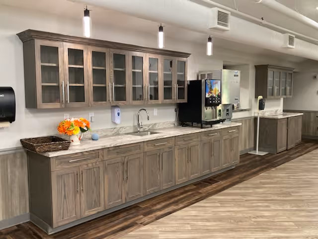 A long countertop with wooden cabinets above and below, featuring a sink, a coffee or beverage machine, a basket with flowers, and a hand sanitizer dispenser mounted on the wall. The room has wood flooring and a white ceiling with exposed ductwork and hanging lights.