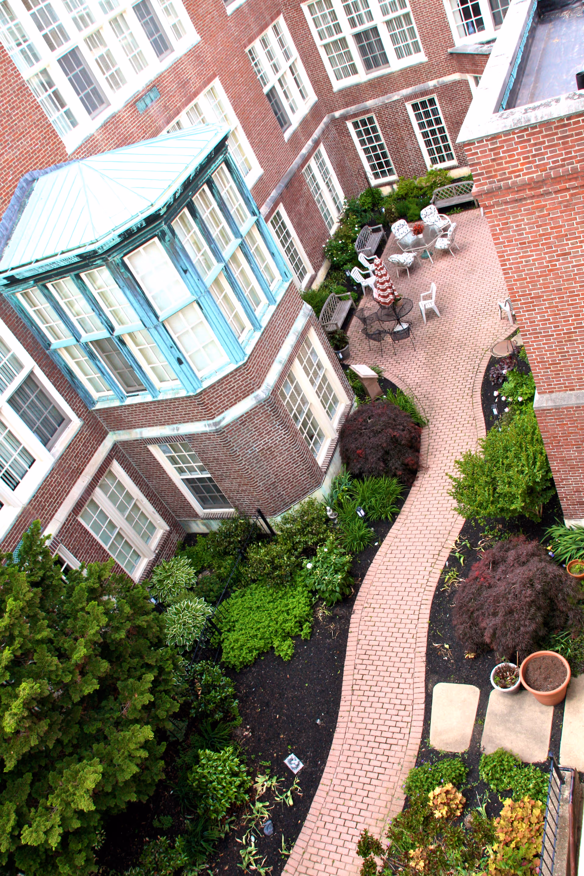 A brick-paved courtyard garden surrounded by a multi-story red brick building with many windows. The courtyard features green shrubs, small trees, and a seating area with white chairs, a table, and a red-and-white striped umbrella.