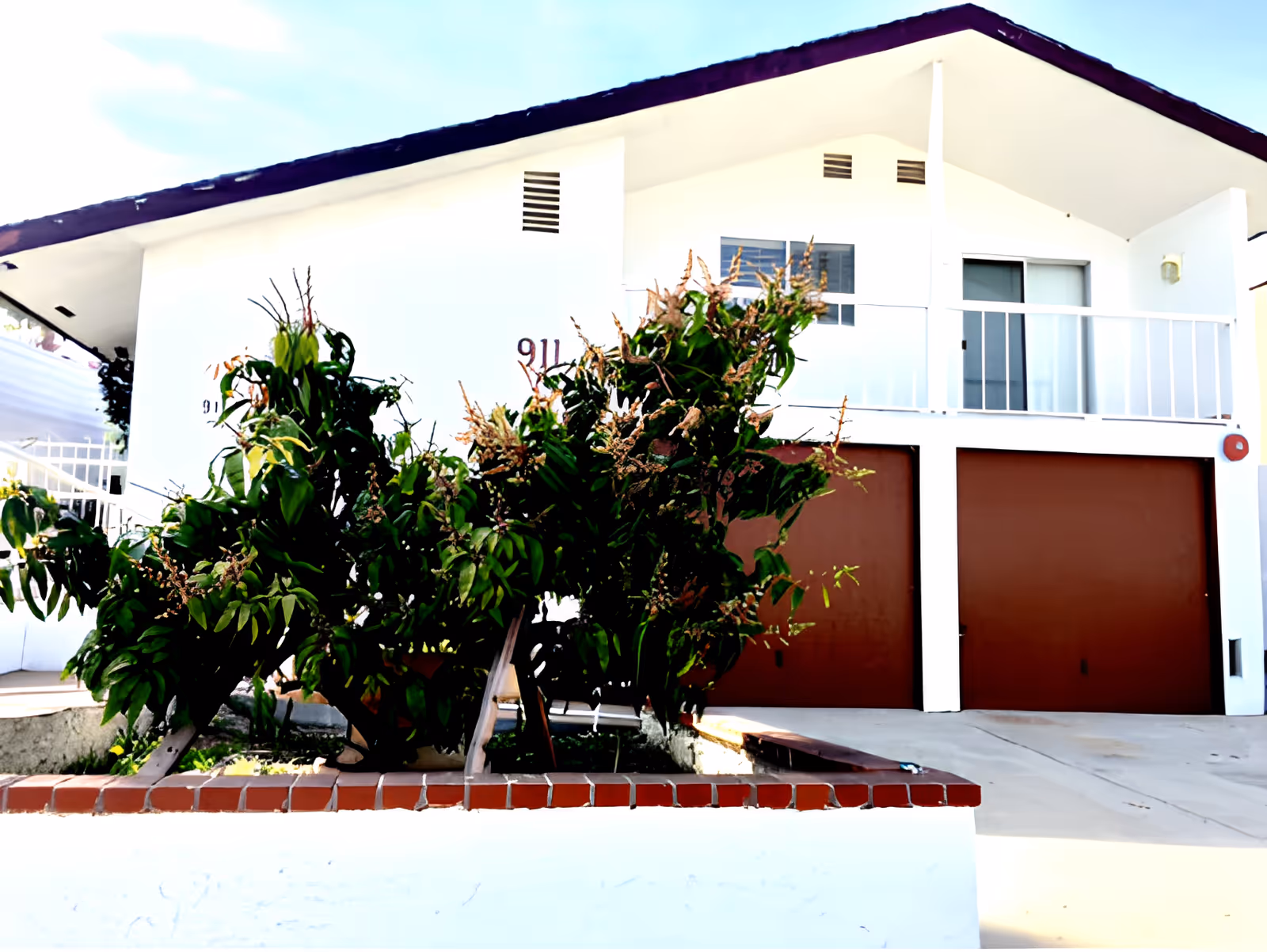 Exterior view of a white two-story building with a slanted roof, featuring two brown garage doors on the ground level and a balcony with a sliding glass door on the upper level. In front of the building, there is a raised garden bed with green leafy plants. The building number 911 is visible on the wall.